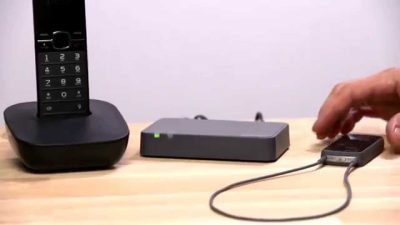 A cordless phone rests on its base beside an Oticon ConnectLine Phone adapter 2.0, which is wired to a smartphone, as a hand reaches for the phone—all placed on a light wooden surface.