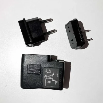 Three black ComPilot Power Supply plug adapters rest on a white surface. Two have different prongs (European and American), while one base unit shows a diagram for attaching the adapters.