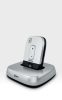 A small, silver Widex TV-DEX wireless device with buttons stands upright in its matching square charging base, both showing the Widex logo, against a plain light background.